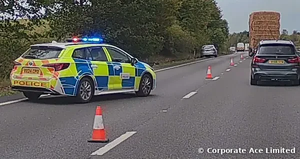 Police car with blue lights at an incident on an A-road, with traffic cones and a lorry carrying hay bales